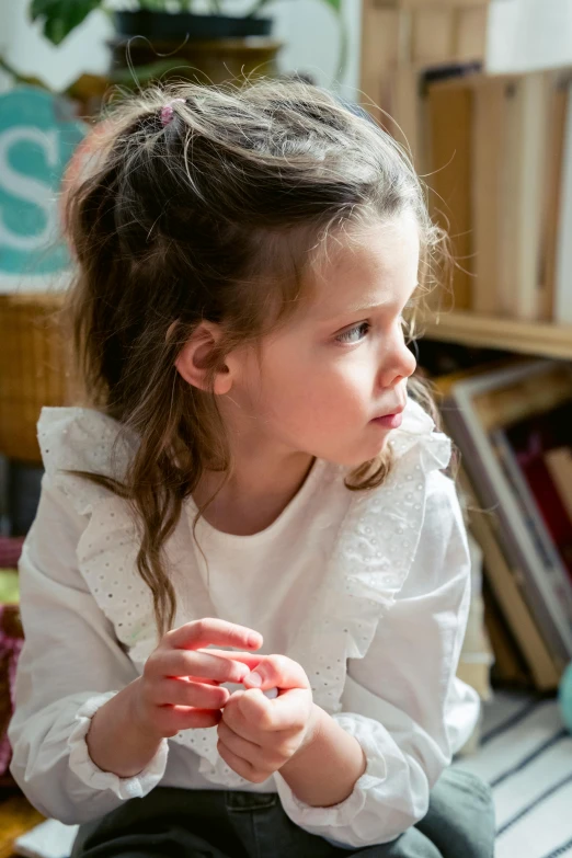 a little girl sitting on top of a wooden floor, inspired by Elsa Beskow, pexels contest winner, pensive expression, white ribbon, looking across the shoulder, gif