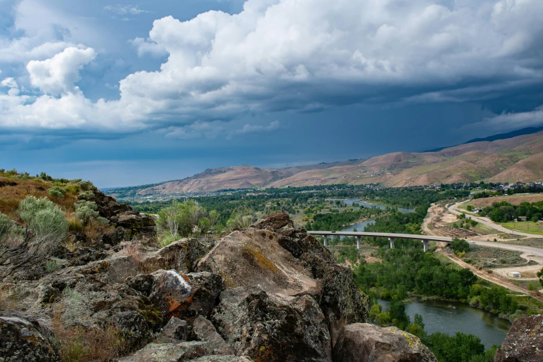 a man standing on top of a rock next to a river, by Arnie Swekel, unsplash contest winner, storm clouds in the distance, idaho, bridge, distant town in valley and hills