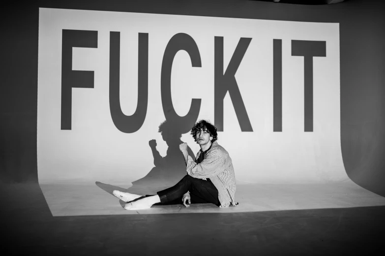 a man sitting on the ground in front of a sign, inspired by Tracey Emin, fluxus, finn wolfhard, sexy pudica pose gesture, in black and white, riccardo scamarcio