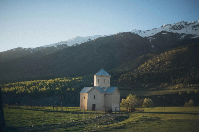 a small white church sitting on top of a lush green field, by Muggur, unsplash contest winner, hurufiyya, snow capped mountains, 1990's photo, late afternoon light, located in hajibektash complex