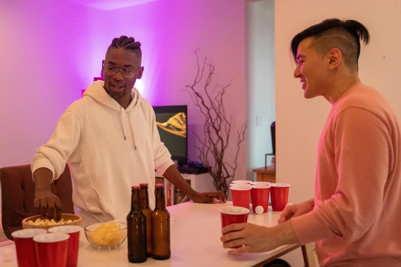 two men sitting at a table with beer and snacks, by Carey Morris, happening, brightly lit pink room, ross tran and bayard wu, airbnb, “house music rave with dancers