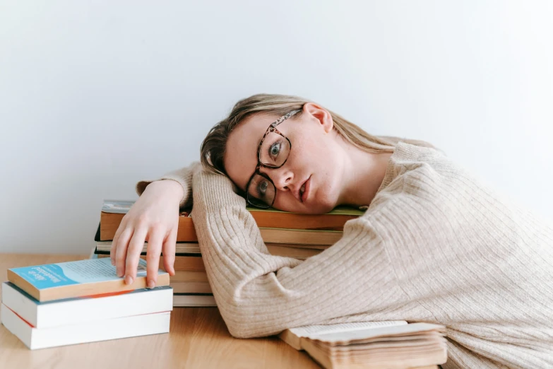 a woman laying her head on a stack of books, a portrait, trending on pexels, with square glasses, exasperated, sydney sweeney, school curriculum expert