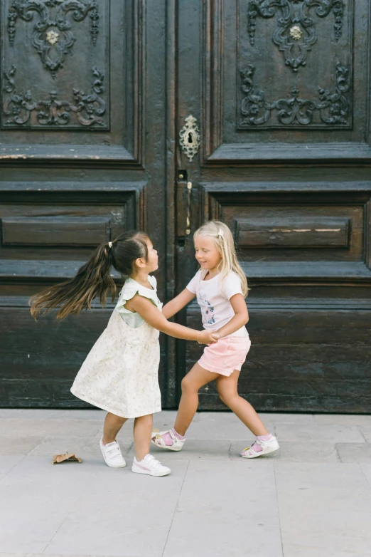 two little girls playing with each other in front of a wooden door, by Nina Hamnett, pexels contest winner, happening, palace dance, in a square, panoramic view of girl, 15081959 21121991 01012000 4k
