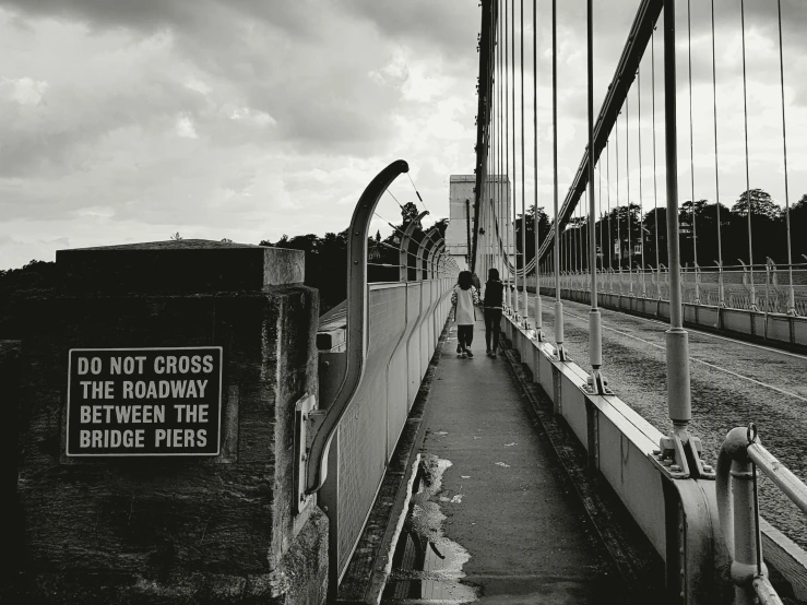 a bridge with a sign that says don't cross the road between the bridge piers, a black and white photo, unsplash, graffiti, people at work, looking at the ground, matt rhodes, from the distance