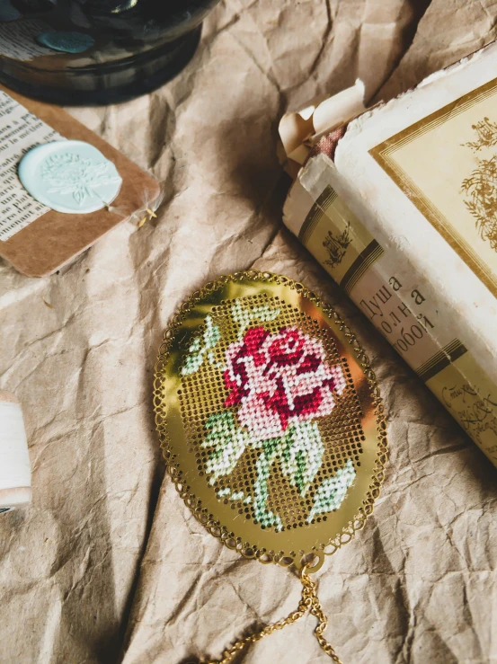 a book sitting on top of a table next to a glass, a cross stitch, inspired by Annie Rose Laing, trending on unsplash, large rose flower head, golden etched breastplate, vintage postcard, closeup - view