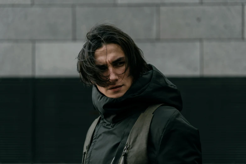 a man standing in front of a brick wall, a character portrait, pexels contest winner, model wears a puffer jacket, kylo ren, frank dillane, messy shoulder-length dark hair