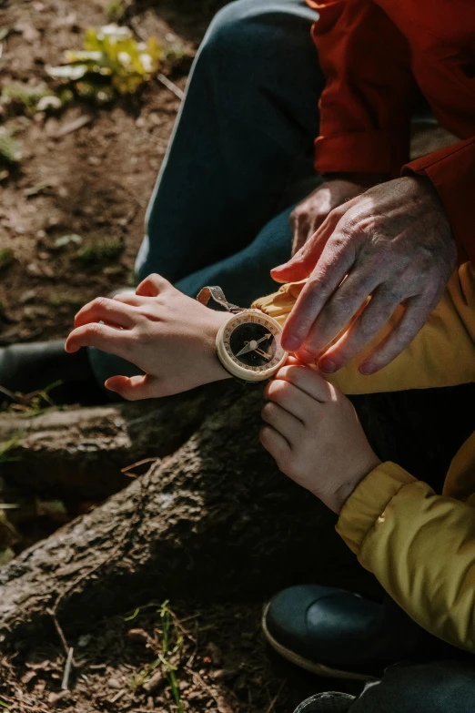 a couple of people sitting next to each other near a tree, wearing a watch, holding a wood piece, adventure gear, family friendly