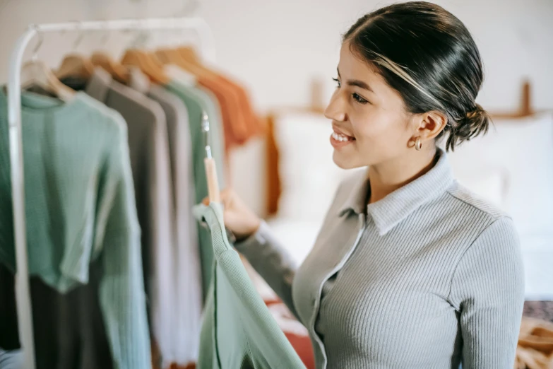 a woman standing in front of a rack of clothes, inspect in inventory image, warm friendly expression, green and brown clothes, thumbnail