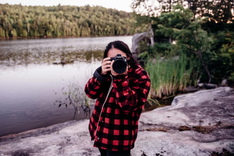 a woman taking a picture of a lake with a camera, by Julia Pishtar, lumberjack flannel, black and red jacket, on a canva, looking up at camera