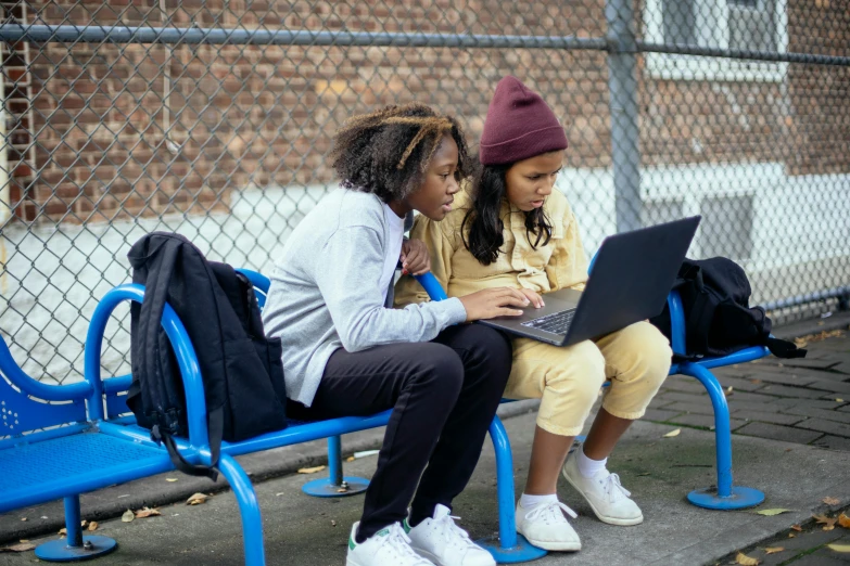 a couple of women sitting on top of a blue bench, trending on pexels, creative coder with a computer, school class, urban playground, sheltered