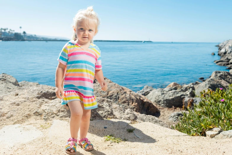 a little girl standing on top of a sandy beach, colourful clothing, tiny feet, stripes, on a rock