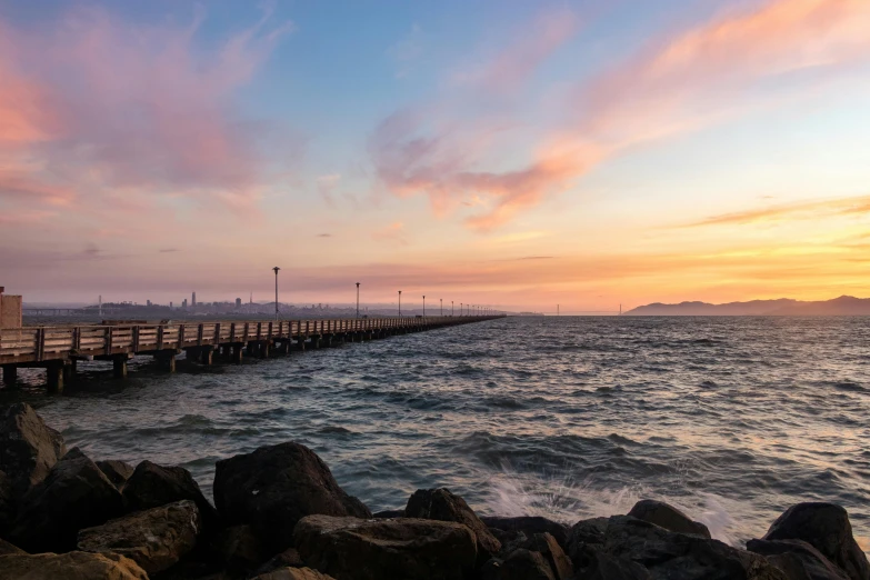 a pier that is next to a body of water, pexels contest winner, pastel sunset, city in the distance, lachlan bailey, a photo of the ocean