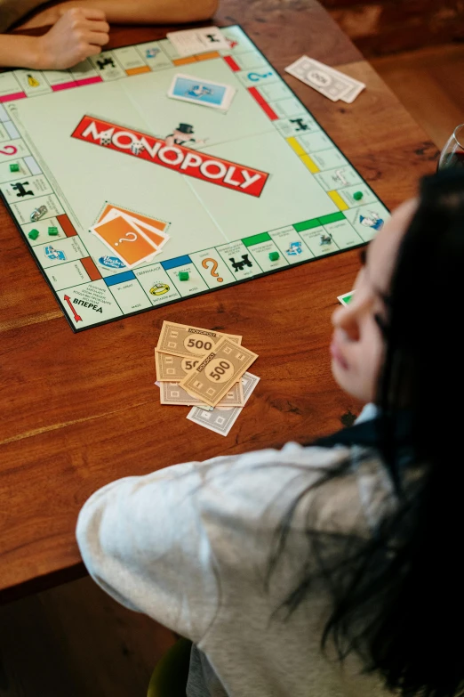 a couple of people sitting at a table playing monopoly, a cartoon, unsplash, visual art, teenage girl, on wooden table, facing away, kid