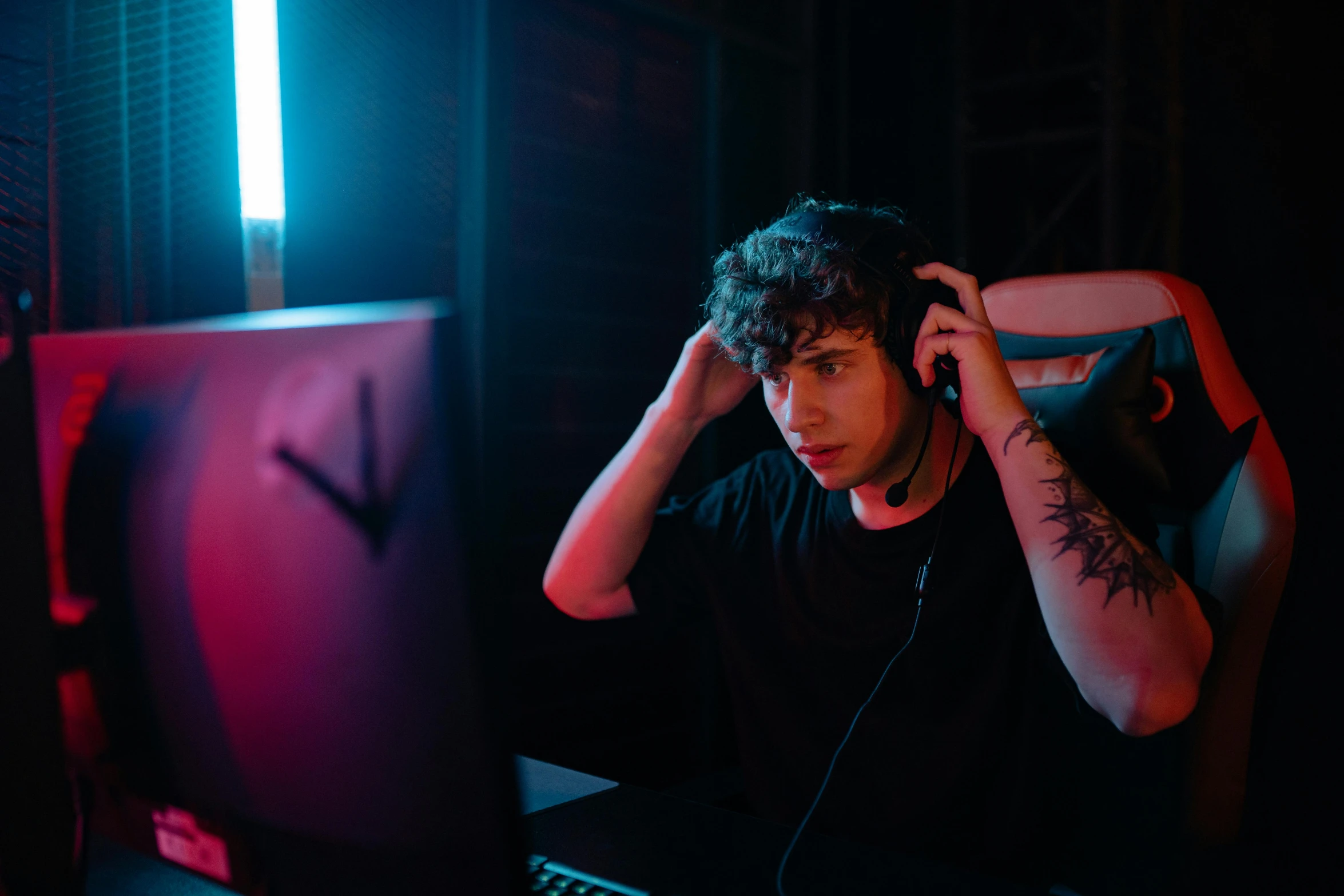 a man sitting in front of a computer in a dark room, an album cover, inspired by John Luke, trending on pexels, realism, gaming headset, curls on top of his head, bisexual lighting, in an action pose