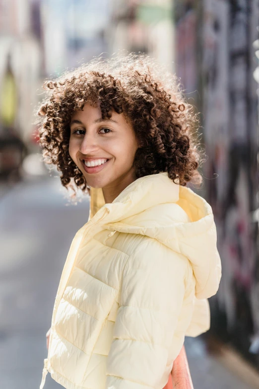 a woman with curly hair standing on a city street, trending on pexels, model wears a puffer jacket, slight yellow hue, portrait of happy a young woman, full frame image