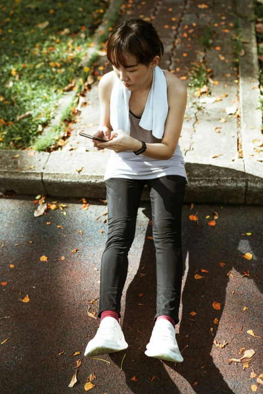 a woman sitting on a curb using a cell phone, trending on pexels, happening, wearing fitness gear, sitting on a leaf, standing, looking down