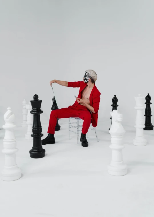 a man sitting on a chair surrounded by chess pieces, an album cover, by Emma Andijewska, trending on pexels, red suit, xqc, zentai suit, official music video