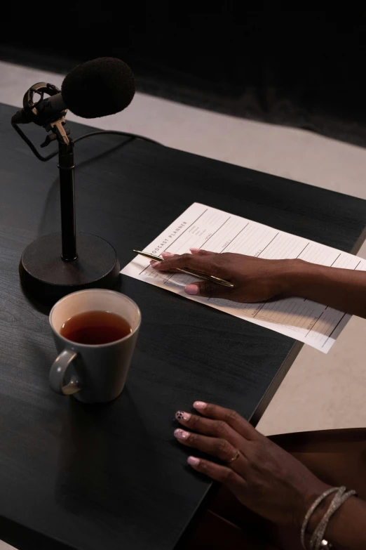 a woman sitting at a table with a cup of coffee, a list cast, live broadcast, curated collections, pen and paper