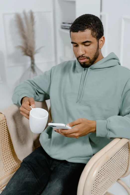 a man sitting on a chair holding a cup and a cell phone, light grey hoodie opened, seafoam green, with a white mug, curated collections