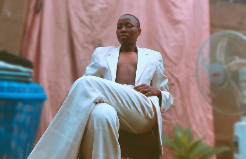 a man sitting on a chair in front of a fan, an album cover, by Emma Andijewska, trending on pexels, lupita nyong'o, wearing white suit, portrait androgynous girl, brown skin