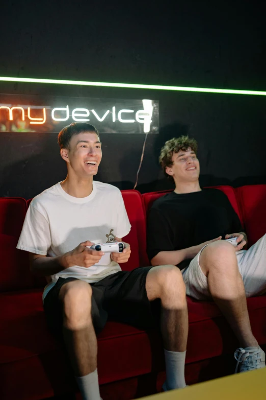 a couple of men sitting on top of a red couch, holding controller, lee madgwick & liam wong, server in the middle, physical : tinyest midriff ever