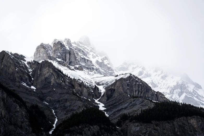 a large mountain covered in snow next to a forest, pexels contest winner, under a gray foggy sky, banff national park, rugged face, high textures