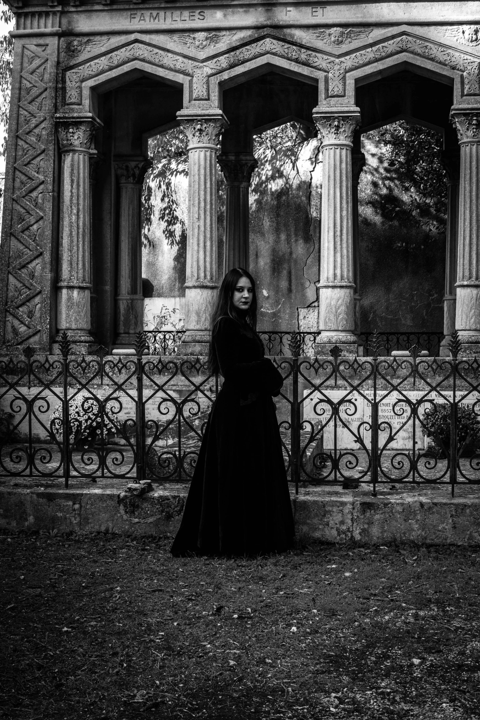 a woman in a black dress standing in front of a building, a black and white photo, pexels contest winner, gothic art, grave, black robes, black metal rococo, early evening