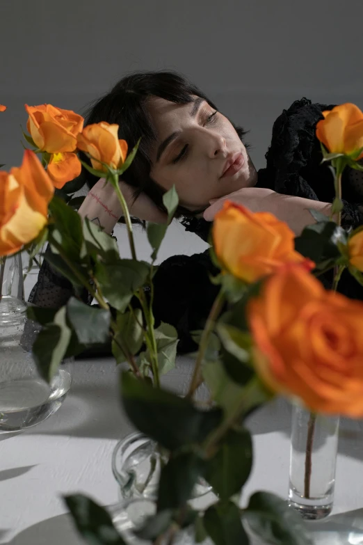 a woman sitting at a table with flowers in front of her, an album cover, trending on pexels, romanticism, black and orange, a pale skin, laying on roses, androgynous person