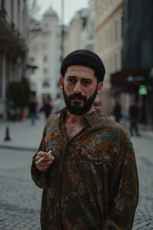 a man standing on a city street smoking a cigarette, an album cover, inspired by Nadim Karam, pexels contest winner, curled beard, hasan piker, vintage color, brown shirt