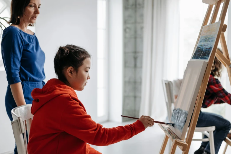 a woman sitting on a chair in front of a painting easel, pexels contest winner, kid drawing, a painting of two people, youtube thumbnail, on artstastion