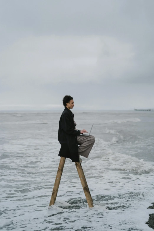 a man sitting on top of a wooden ladder next to the ocean, unsplash, conceptual art, woman in black robes, sitting at a computer, on a cloudy day, on stilts