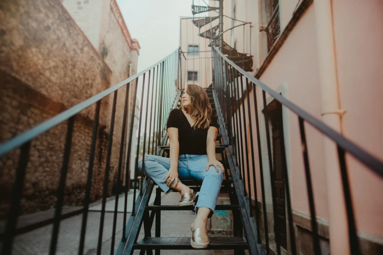 a woman sitting on the steps of a building, by Emma Andijewska, pexels contest winner, wearing blue jean overalls, alley, background image, looking up at the camera