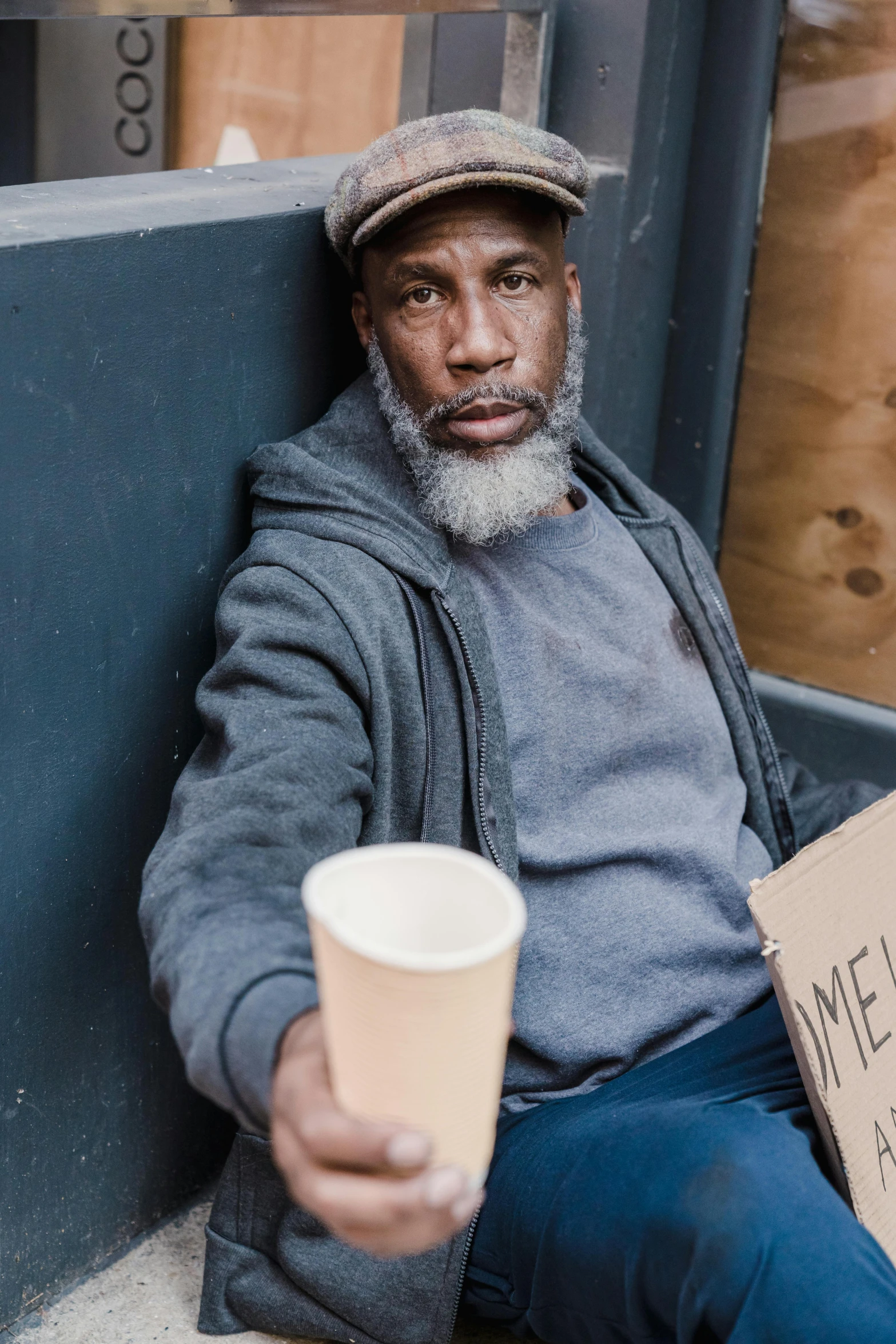 a man sitting on the sidewalk holding a cup of coffee, a photo, by Will Ellis, renaissance, malnourished, gray men, promo image, jemal shabazz