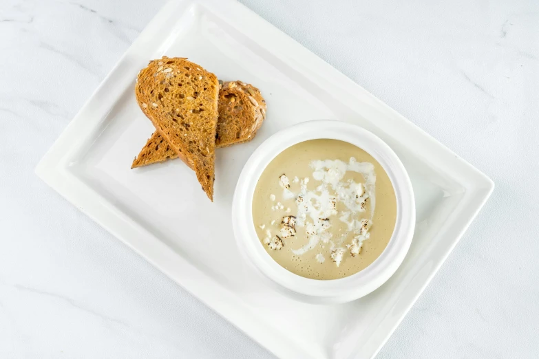 a white plate topped with a bowl of soup next to a piece of bread, taupe, square, productphoto, ballard