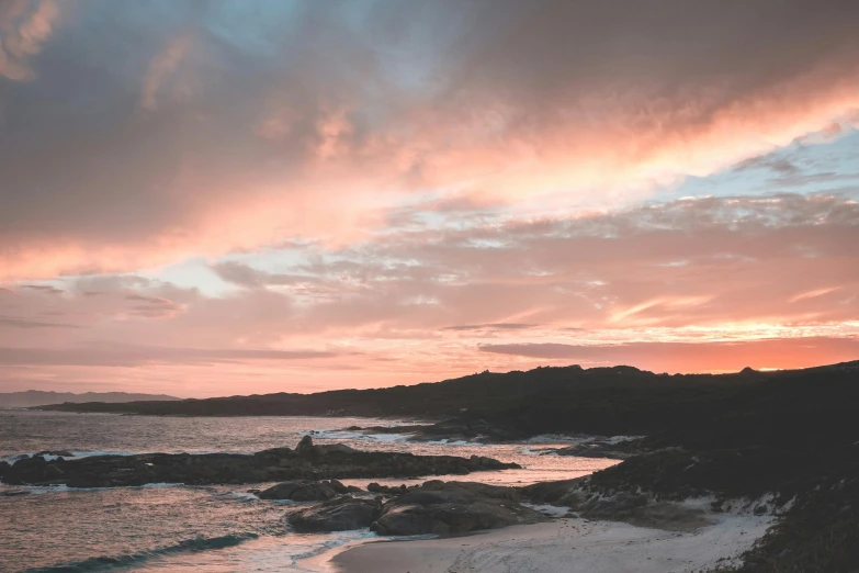 a man standing on top of a sandy beach next to the ocean, by Charlotte Harding, pexels contest winner, romanticism, pink storm clouds, sunset panorama, rocky shore, pink white turquoise