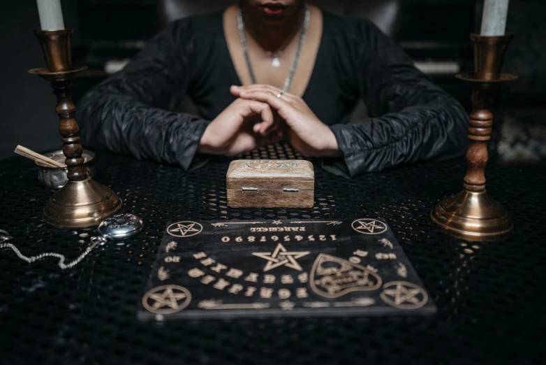a woman sitting at a table with candles in front of her, an etching, trending on pexels, holography, occult robes, game board, a man wearing a black jacket, holding a wood piece
