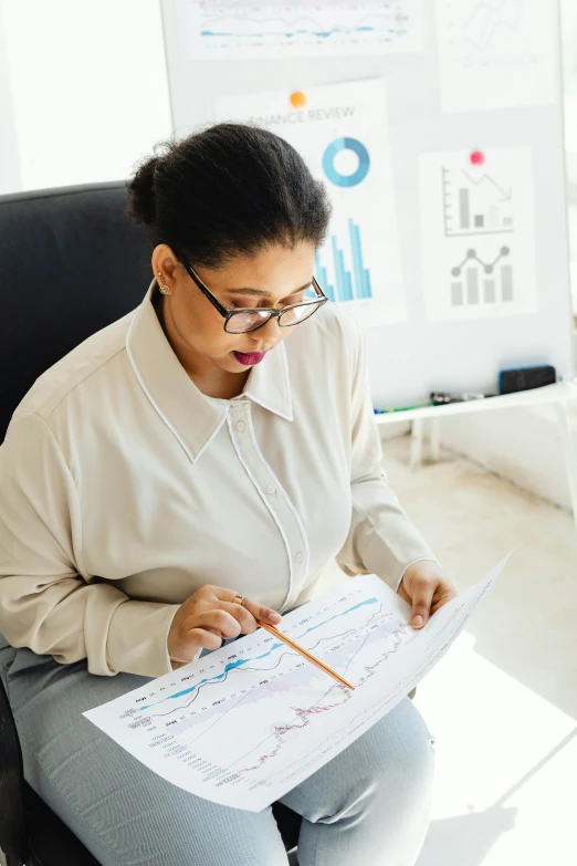 a woman sitting in a chair holding a piece of paper, a cartoon, analytical art, informative graphs and diagrams, sat at her desk, commercially ready, wearing thin large round glasses