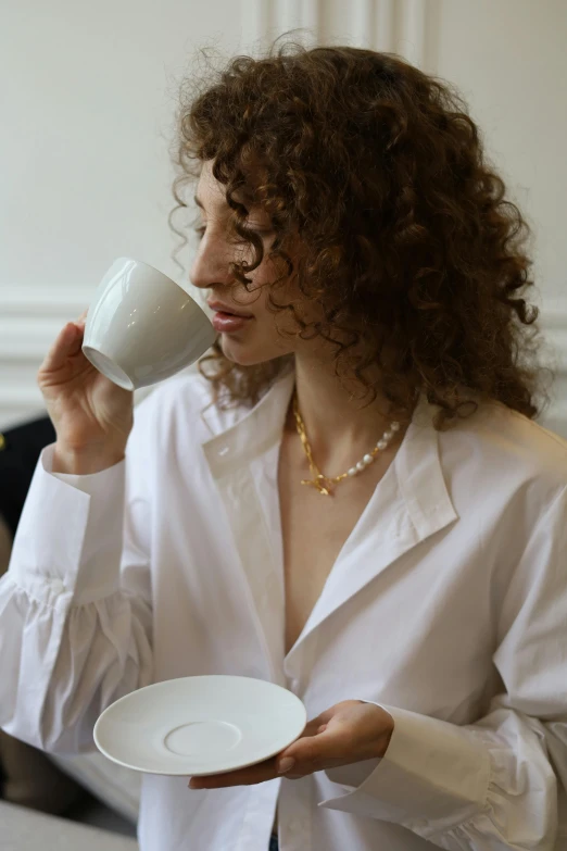 a woman in a white shirt drinking out of a cup, inspired by Mathieu Le Nain, wearing gold jewellery, puka shell necklace, cereal, curated collections