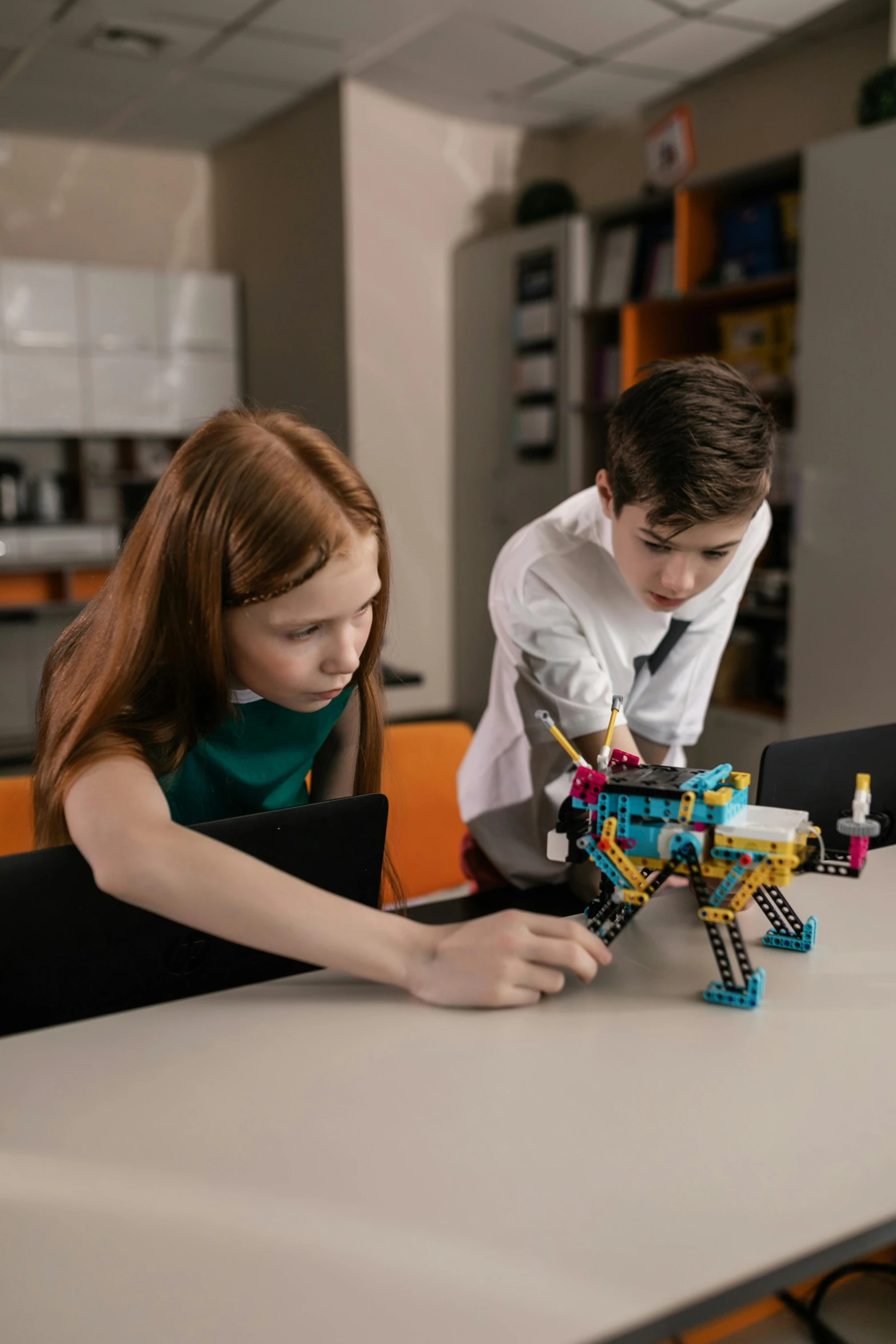 a couple of kids that are sitting at a table, mechanical arm, lego set, back towards camera, robot lion