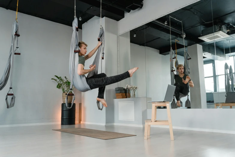 a man flying through the air while riding a hammock, by Emma Andijewska, hurufiyya, dim dingy gym, two hang, reflecting, in a white boho style studio
