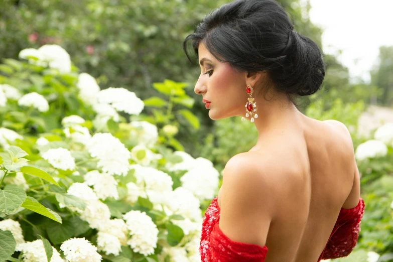 a woman in a red dress standing in front of flowers, photoshoot for skincare brand, showing her shoulder from back, beautiful himalayan woman, wearing ornate earrings