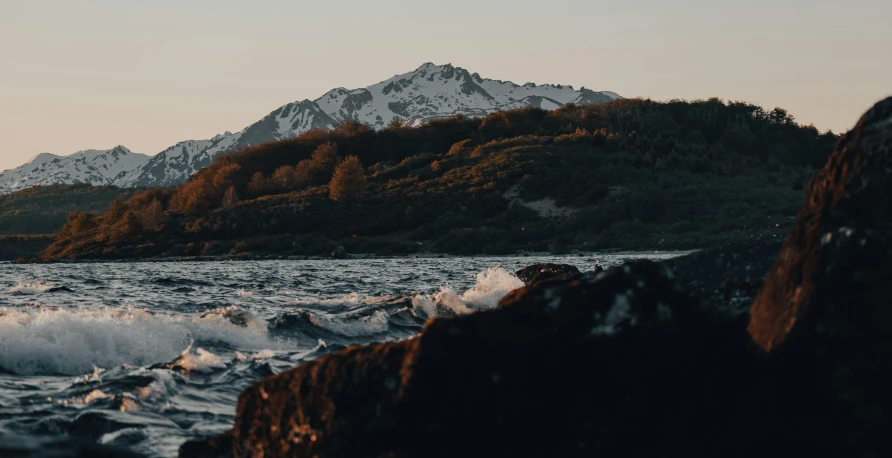 a body of water with a mountain in the background, a picture, unsplash contest winner, romanticism, sea waves, late summer evening, brown, snow capped mountains