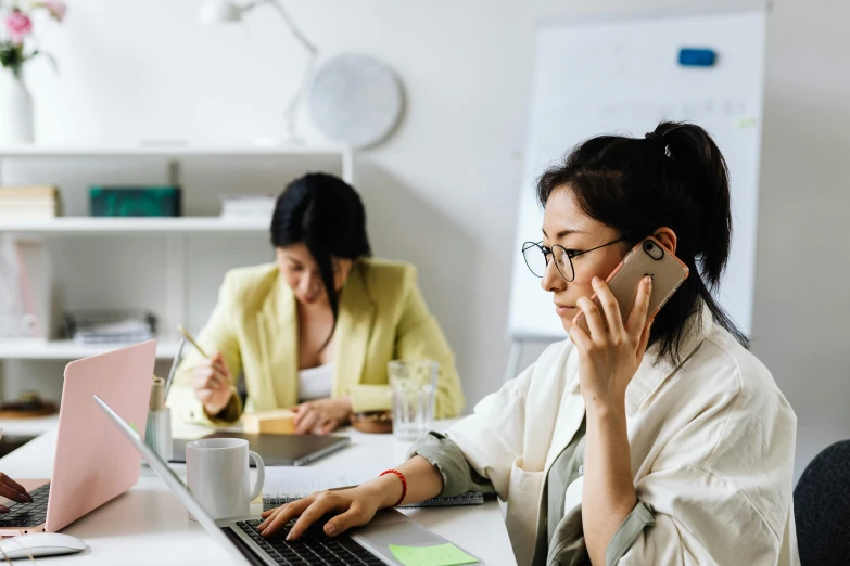 a woman talking on a cell phone while sitting at a desk, by Lee Loughridge, trending on pexels, hurufiyya, two women, in an call centre office, asian human, working on a laptop at a desk