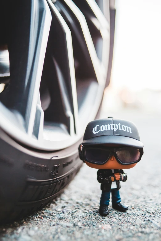 a close up of a toy in front of a tire, a cartoon, inspired by An Gyeon, unsplash contest winner, graffiti, wearing sunglasses and a cap, compulsion, standing in front of lowrider, funko pop