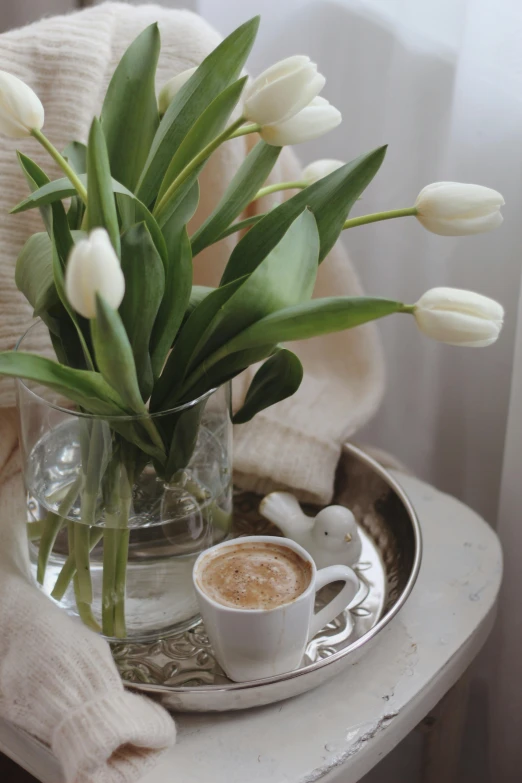 a glass vase filled with white tulips next to a cup of coffee, inspired by Judy Takács, romanticism, silver, cottagecore, milk and mocha style, highly polished