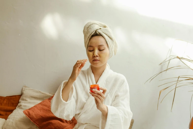 a woman sitting on a bed with a towel on her head, a picture, trending on pexels, having a snack, orange robe, korean face features, partially cupping her hands