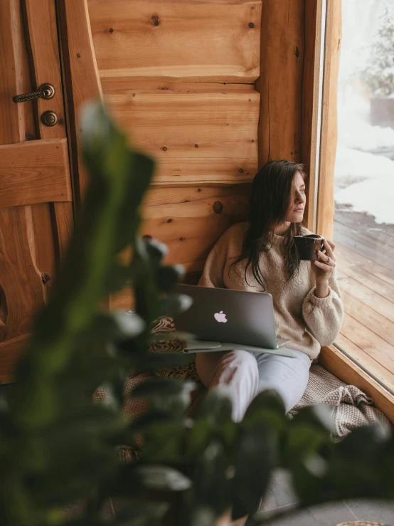 a woman sitting in front of a window with a laptop, by Julia Pishtar, trending on pexels, inside of a cabin, chilling 4 k, next to a plant, profile image