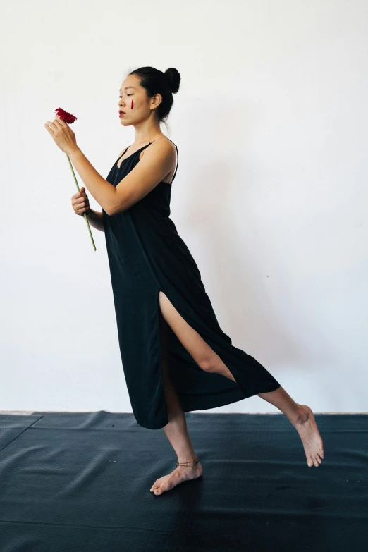 a woman in a black dress holding a red rose, inspired by Ruth Jên, arabesque, standing athletic pose, barefoot in sandals, wearing a long flowy fabric, she is wearing a black tank top