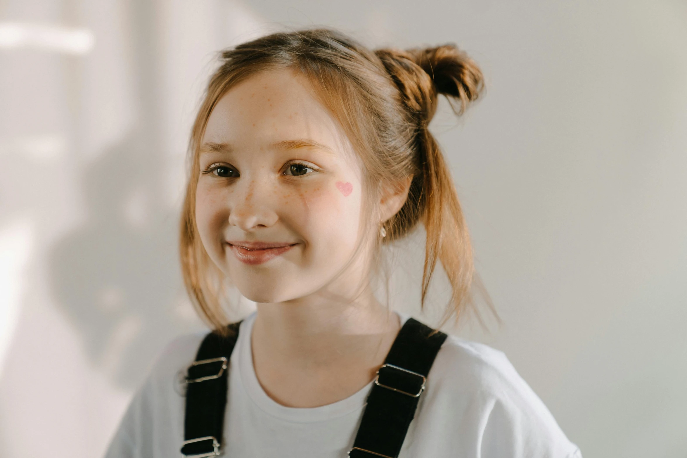 a young girl standing in front of a white wall, a character portrait, pexels contest winner, brown hair in a ponytail, kind smile, trending photo, markings on her face