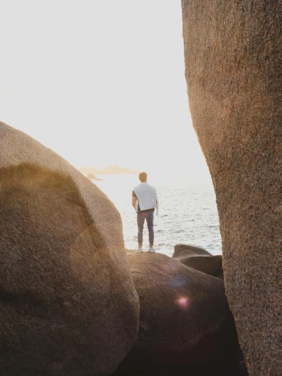 a man standing on top of a large rock next to the ocean, pexels contest winner, looking outside, boulders, slightly pixelated, back lit